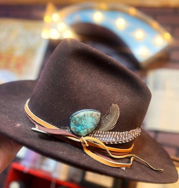 Brown hat with decorative elements including a turquoise stone and feathers, held against a blurred background.