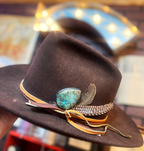 Brown hat with decorative elements including a turquoise stone and feathers, held against a blurred background.