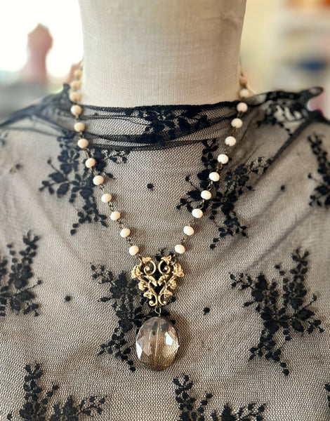 A double-strand necklace displayed on a mannequin torso, featuring small cream-colored beads, an antique gold filigree heart-shaped pendant, and a faceted smoky brown glass drop.