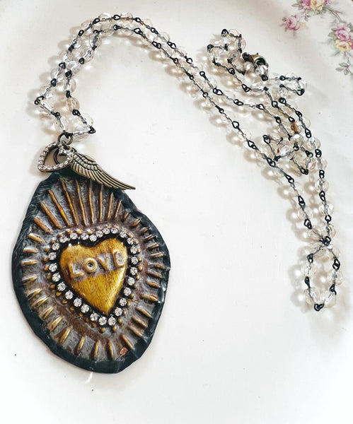 A handmade-style pendant necklace featuring a dark, textured teardrop-shaped charm with a gold heart in the center that reads 'LOVE' and is surrounded by small rhinestones. The pendant is attached to a silver-toned wing charm and a chain mixed with clear faceted beads and black seed beads, displayed on a white floral plate.