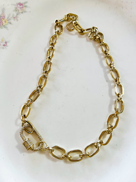 A gold-toned chain link bracelet with a lobster clasp, photographed on a white surface with a faint floral pattern in the upper left corner.