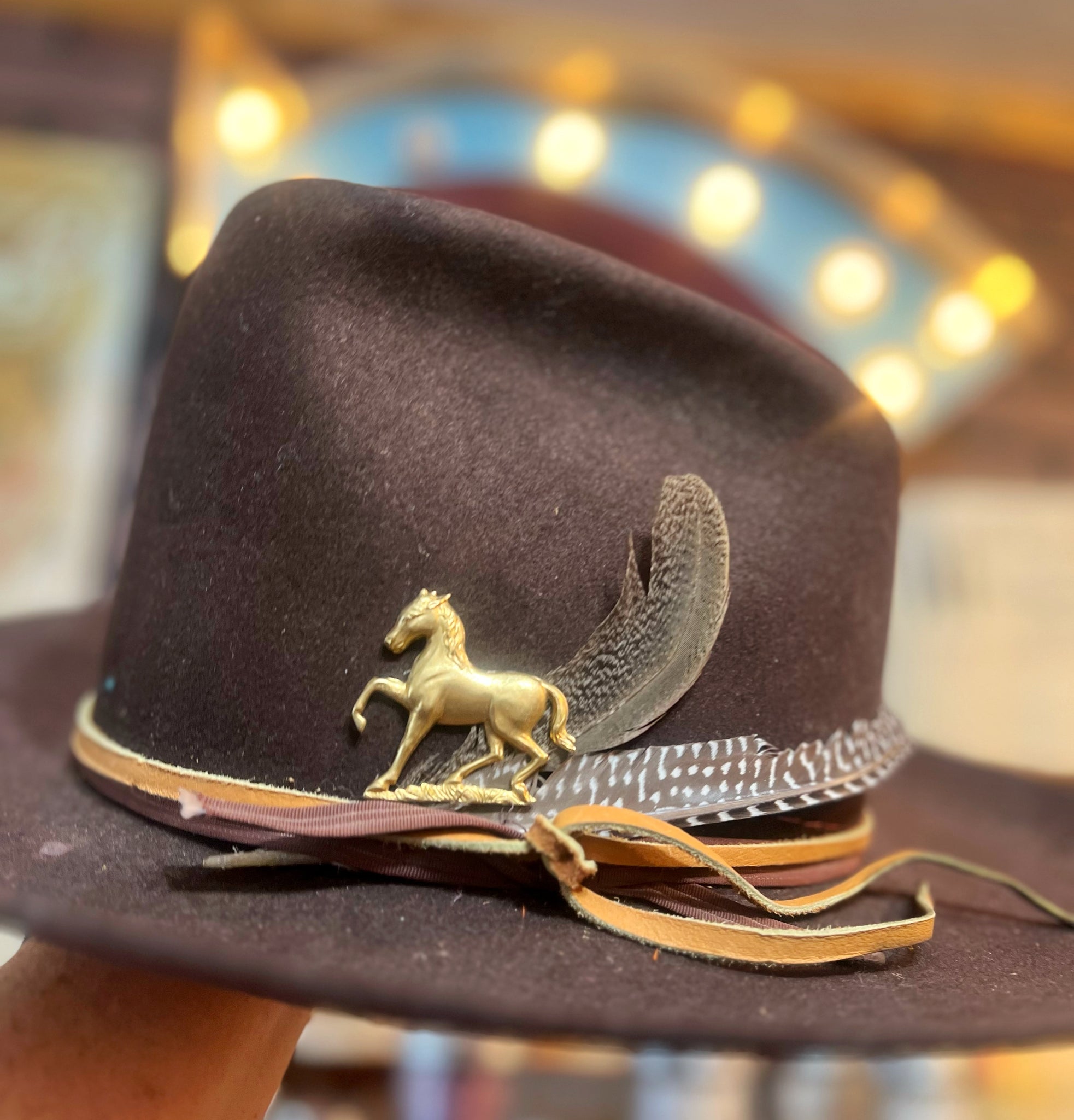 Brown cowboy hat with gold horse emblem and feather detail, blurred background