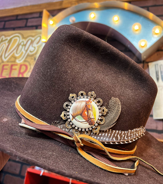 Brown cowboy hat with decorative band featuring a horse emblem, against a vintage-style neon sign background.