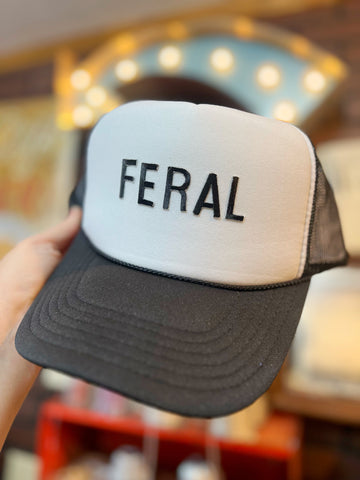 Trucker hat with 'FERAL' text held in front of a blurred indoor background